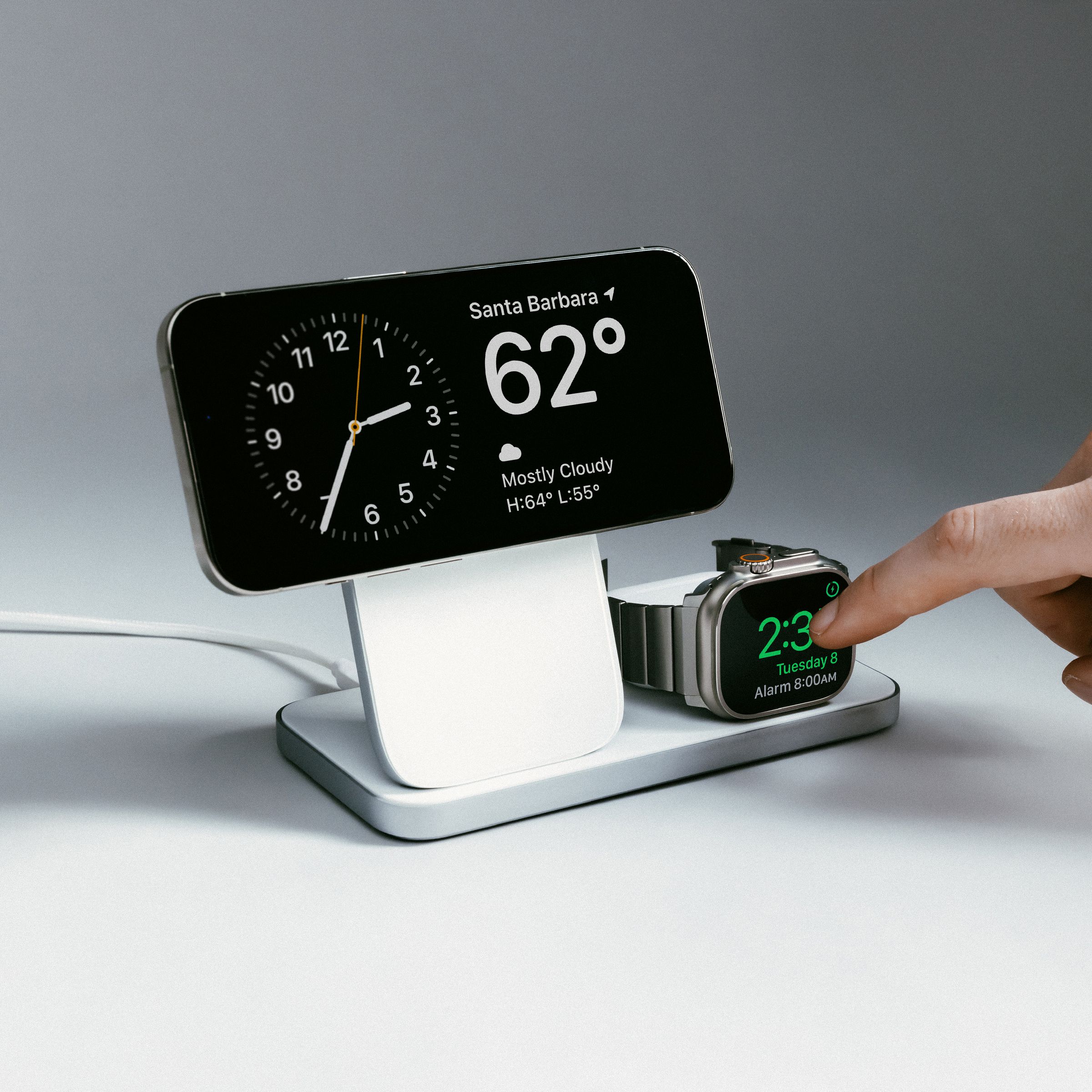 An iPhone and Apple Watch wirelessly charging on a Nomad Stand One Max three-in-one charger. The iPhone is horizontal in StandBy mode and a person is tapping on the Apple Watch screen.