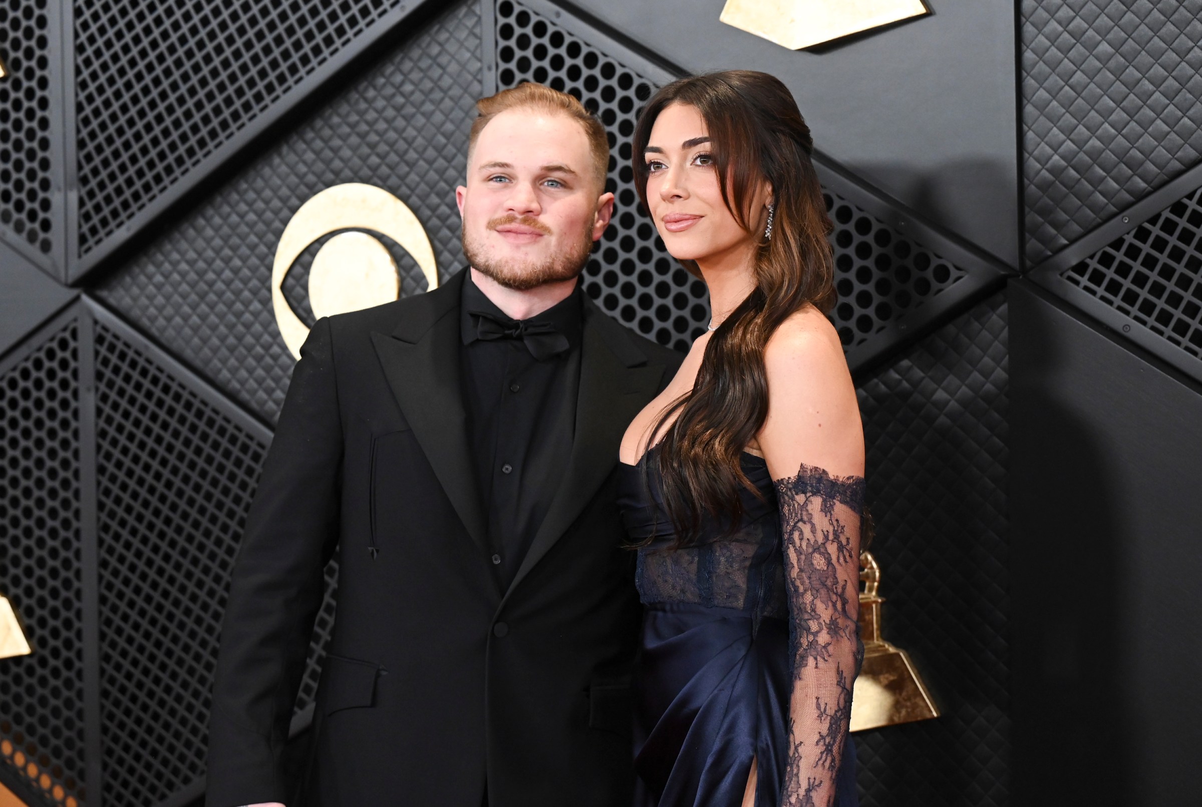 Zach Bryan in an all-black tuxedo posing beside a brunette in a lace corset dress.