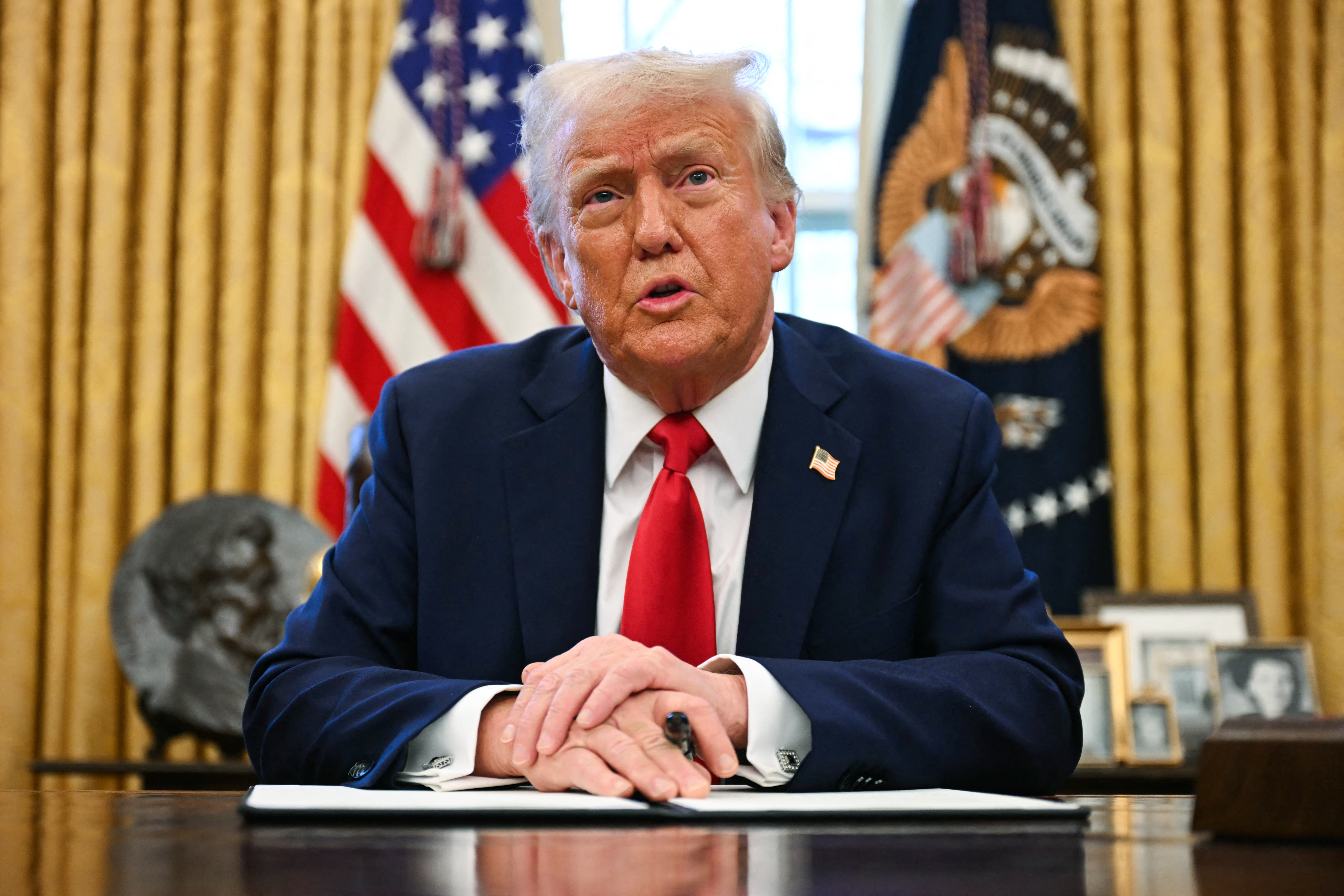 President Trump at his desk, hands folded, a pen in one hand.