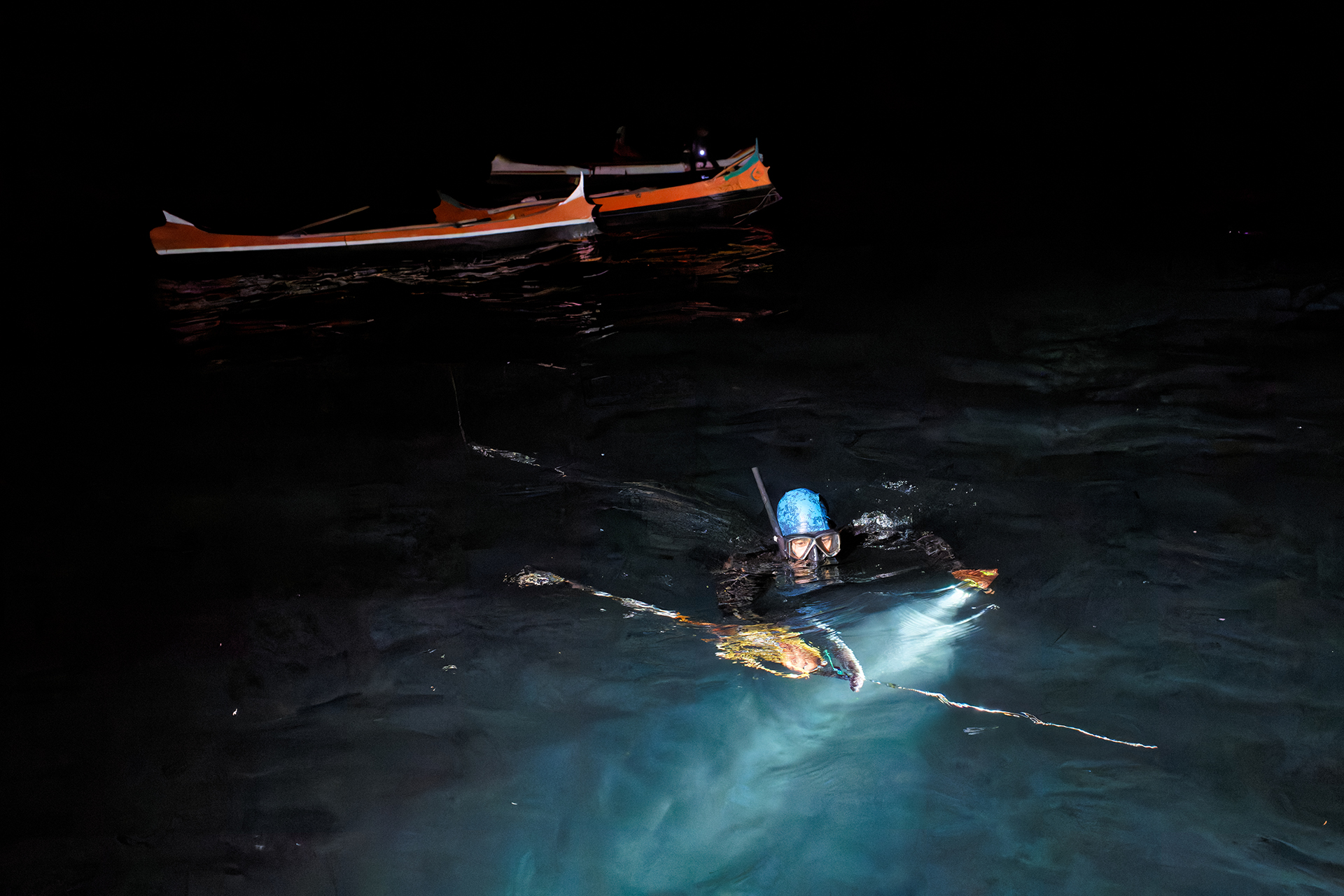 A fisherman with goggles, a spear and underwater flashlight swims near fishing boats at night.