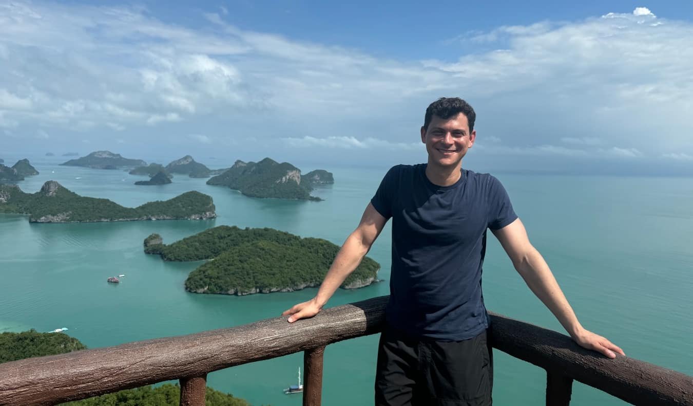 Bedouin Matt in Thailand, smiling and looking at the camera in a stunning view, turquoise waters dotted with green islands behind him