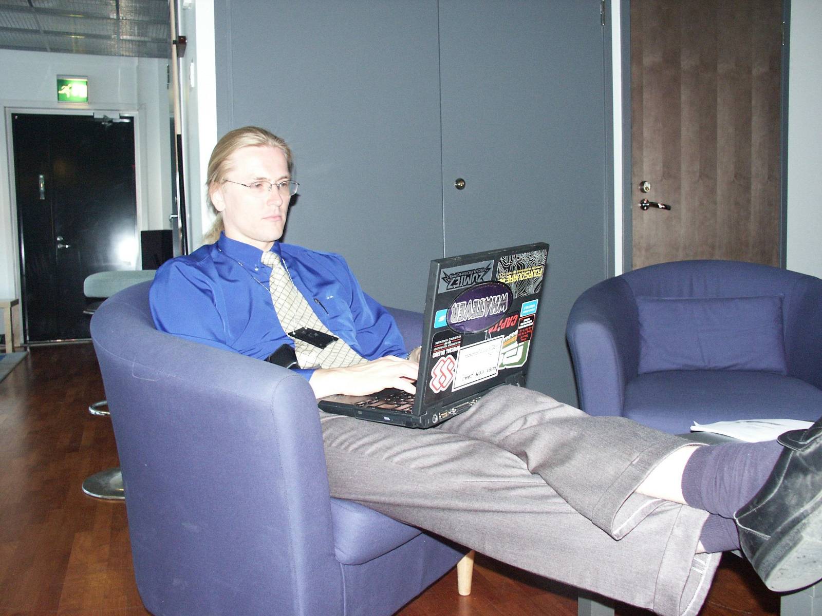Photo of a younger Mikko Hypponen, wearing a blue shirt and tie, in a purple chair, with his feet up and a laptop with stickers on his lap.