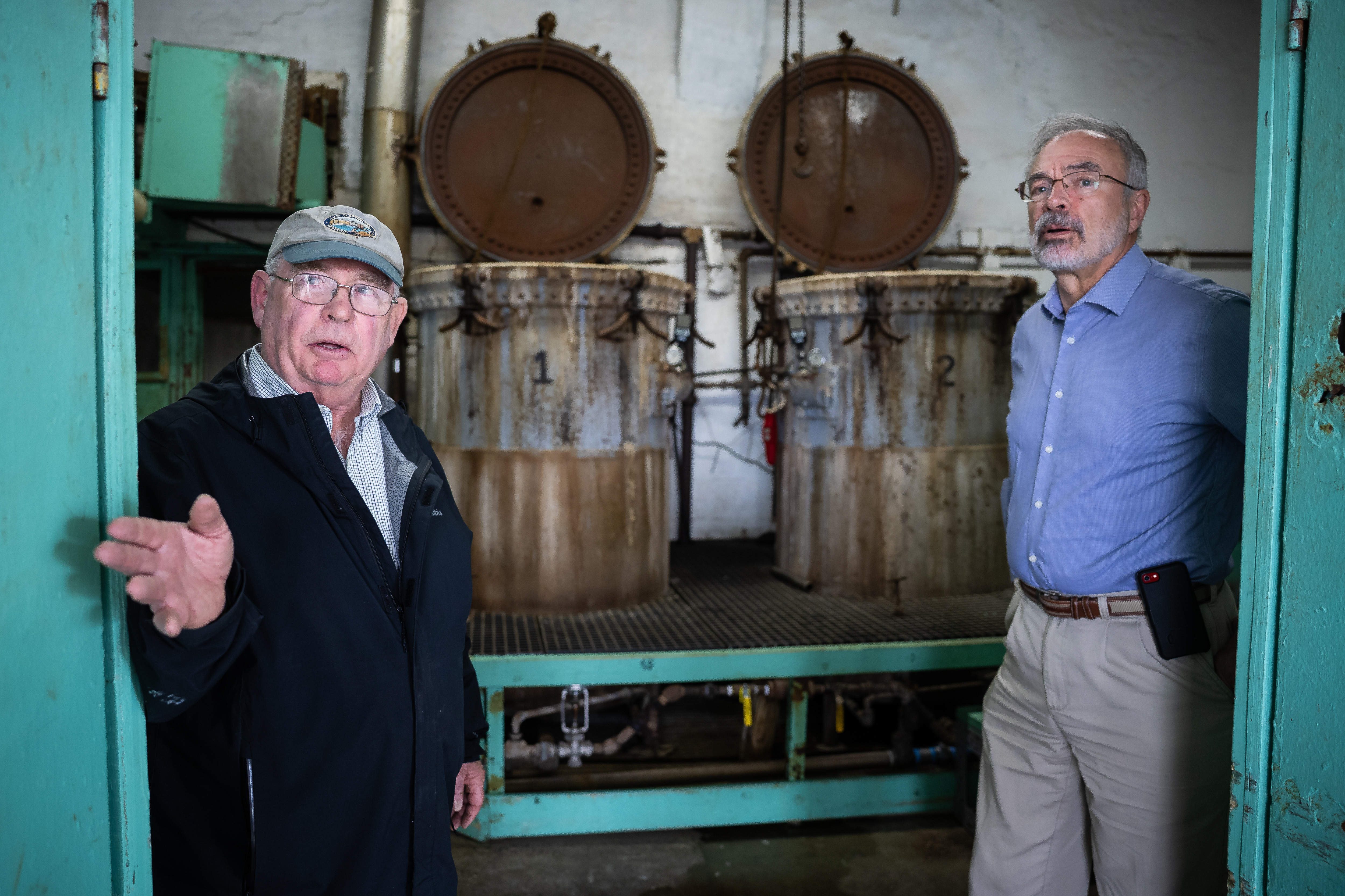 Jack Brooks, owner of the J.M. Clayton Company, speaks with Harris during a tour of the company's crab processing plant. Behind them, crab steamers are seen.