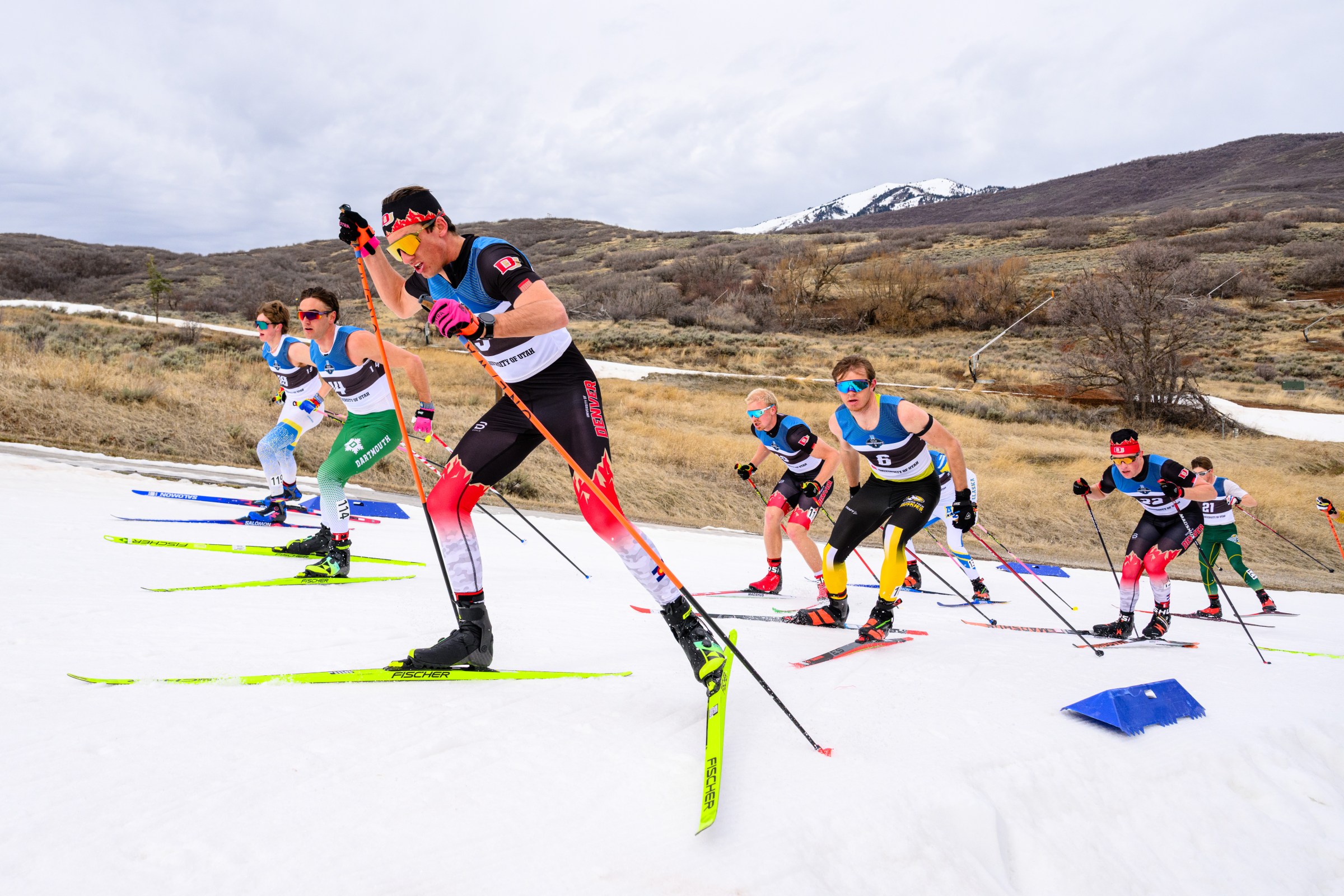 collegiate skiers ski up a narrow path of snow with bare grassy hills around them