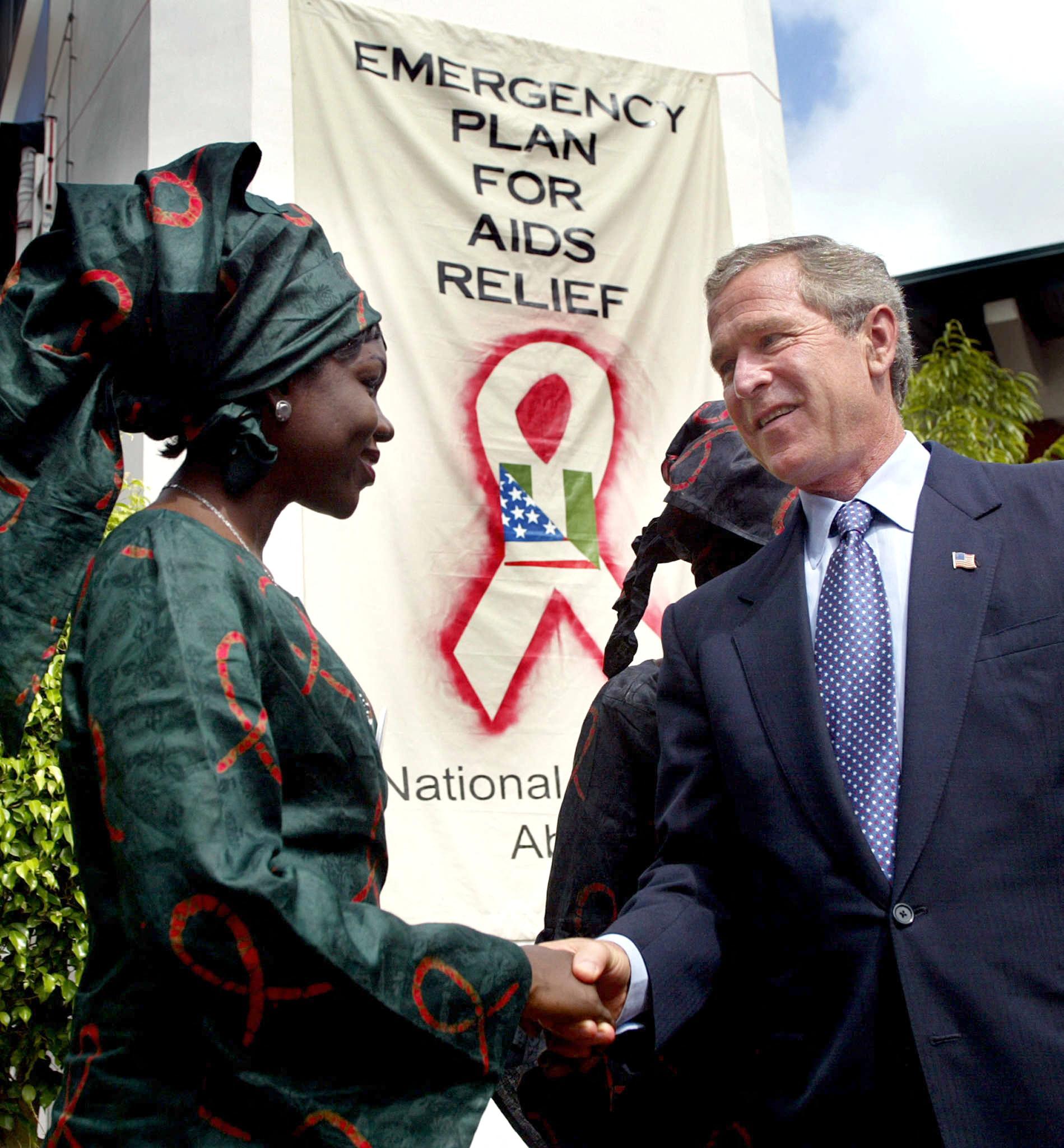 George Bush shakes hands with a woman in front of a sign that reads “Emergency Plan for AIDS Relief”